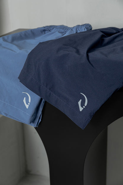 A pair of blue athletic shorts with a white logo on the left leg, displayed on a dark surface.